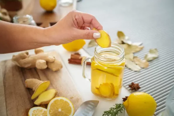 A hand placing fresh ginger slice into a jar of lemon and ginger water, surrounded by raw ginger, lemon, cinnamon, and bay leaves.